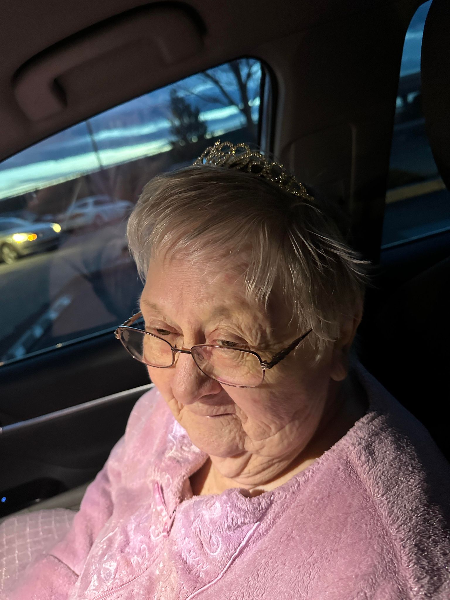 Elderly woman wearing a pink bathrobe in a car with a faux tiara on her head. 
